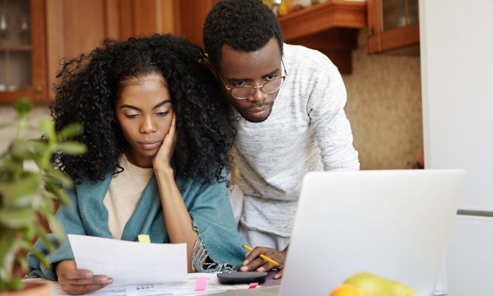 Young African family paying utility bills online using laptop computer. Unhappy female sitting at table analyzing sheet of paper in her hand, calculating domestic expenses together with her husband