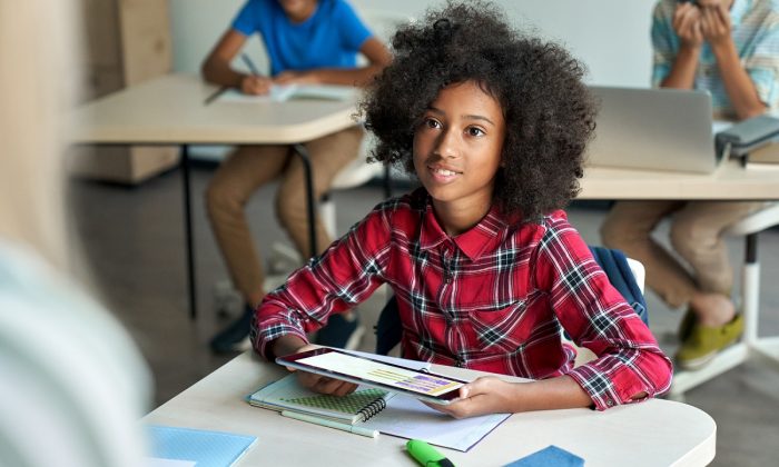 Happy Afro American schoolgirl looking at teacher holding tablet in classroom.