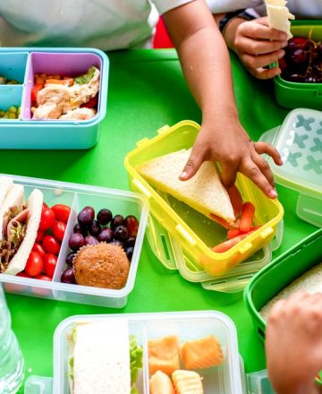 Children's,Hands,Reaching,For,Colorful,Lunchboxes,Filled,With,Sandwiches,,Fruits,
