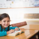 Child,Female,Student,Sitting,In,Classroom,At,Desk,Reading,Book,