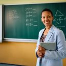 Happy,African,American,Teacher,Standing,In,Front,Of,Chalkboard,At