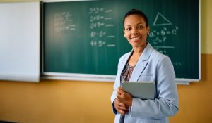 Happy,African,American,Teacher,Standing,In,Front,Of,Chalkboard,At